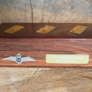 Puede incluir: Una caja de madera con una placa de latón que dice "Su mensaje grabado aquí gratis". La caja tiene un emblema de la Royal Air Force en la parte delantera y tres dominós incrustados en la parte superior.