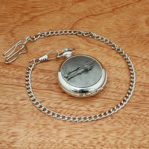 Könnte beinhalten: Eine silberne Taschenuhr mit einer Kette. Das Zifferblatt der Uhr ist mit einer detaillierten Gravur eines Flugzeugs verziert.