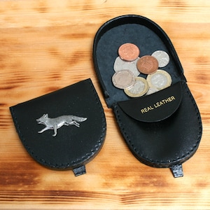 May include: Two black leather coin purses, one open and filled with coins. The open purse has "REAL LEATHER" printed on the flap. The closed purse features a silver fox emblem. Both purses are on a wooden surface.