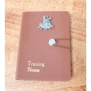 Può includere: Un taccuino in pelle marrone con una figurina argentata di una coppia che balla sul davanti. Il taccuino ha un'iscrizione dorata che recita "Training Notes".