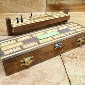 May include: A wooden cribbage board with a hinged lid and brass clasps. The board has a light wood inlay and a Scottish flag on the side. The board is closed and sitting on a wooden surface.