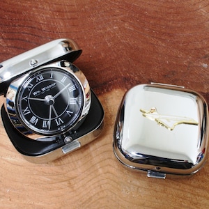 May include: A silver-colored travel clock with a black face and Roman numerals. The clock is open, revealing the time. Next to it is a matching silver case with a gold airplane emblem. The clock face reads "WM. WIDDOP EST. 1987."