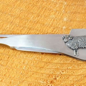 May include: A silver-colored pocket knife with a sheep design on the handle. The knife has a small blade and a tool for packing tobacco.
