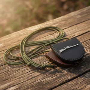 May include: A black leather pouch with a silver rifle emblem, placed on a brown leather pouch. An olive green cord is looped around the pouches. The items are on a weathered wooden surface.