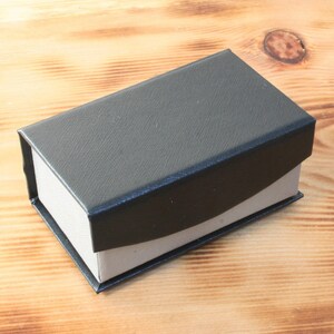 May include: A black rectangular box with a white interior. The box is made of a textured material and has a slightly curved lid.