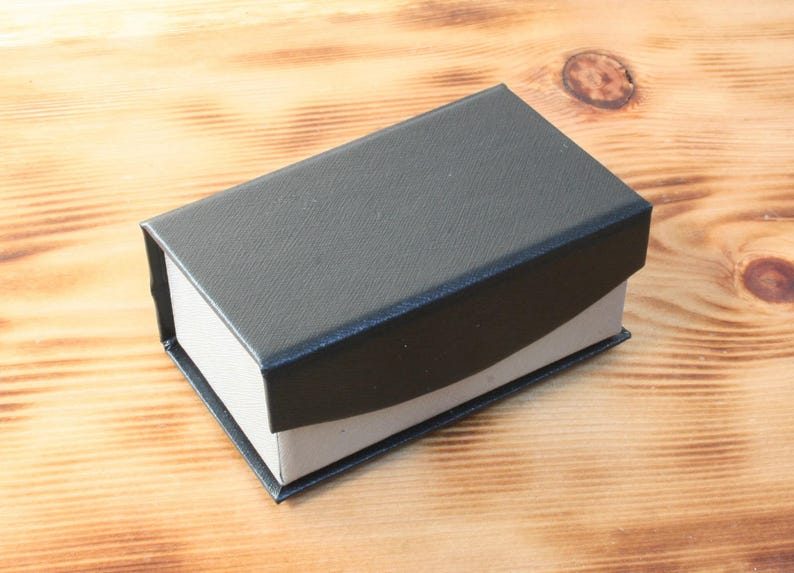 May include: A black rectangular box with a white interior. The box is made of a textured material and has a slightly curved lid.