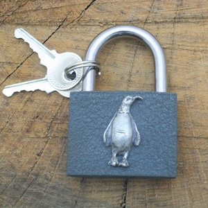 May include: A gray padlock with a silver penguin design. The padlock is attached to a silver key.
