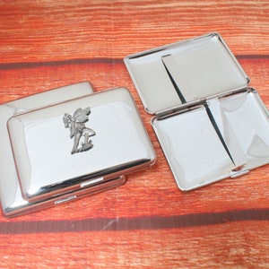 May include: A silver cigarette case with a hinged lid and a small fairy design on the front.