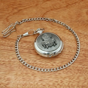 May include: A silver pocket watch with a chain. The watch face features a detailed engraving of a steam locomotive.