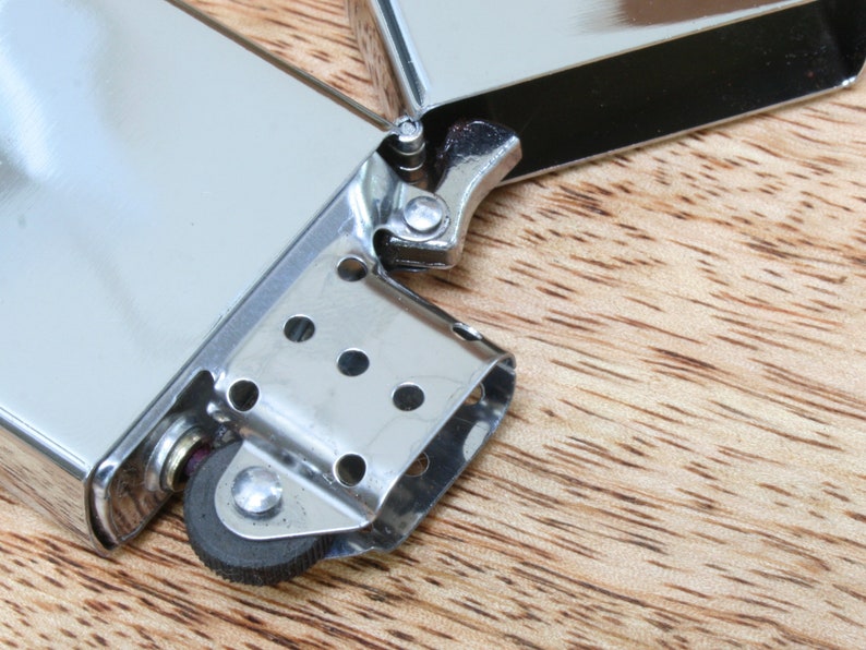 May include: A silver lighter with a black wheel and a silver hinge. The lighter is open and the wheel is visible.