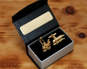 May include: Gold-colored cufflinks in a black and gray gift box. The cufflinks are in the shape of a rocket and a barrel. The box has a gold-colored plate on the inside of the lid and sits on a wooden surface.