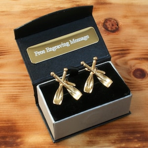 Peut inclure: Boutons de manchette en or dans un coffret noir avec un intérieur argenté. Les boutons de manchette sont en forme de rames croisées. Le coffret porte une étiquette indiquant "Message de gravure gratuit".