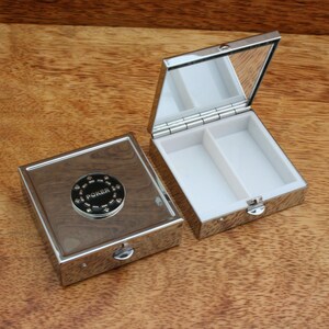 May include: A silver pill box with a wood-grain design and a poker chip on the lid. The box is open and has two compartments inside.