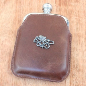 Könnte beinhalten: Eine braune Lederflasche mit einem silbernen Kraken-Design auf der Vorderseite.