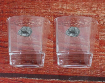 May include: Two clear glass shot glasses with a pewter cowboy hat design on the front.