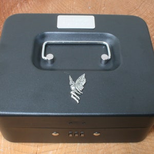 Könnte beinhalten: Eine schwarze Metall-Geldkassette mit einem Zahlenschloss und einem silbernen Feendesign auf dem Deckel. Die Kasse hat einen silbernen Griff.