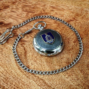 Op de afbeelding: Zilveren zakhorloge met een blauw en gouden wapen op de wijzerplaat en een zilveren ketting.