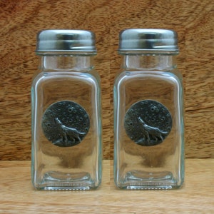 Puede incluir: Dos botes de especias de vidrio transparente con tapas de metal plateado. Cada bote tiene un emblema circular de color peltre con un lobo aullando frente a una luna. Los botes son cuadrados y tienen un diseño clásico.
