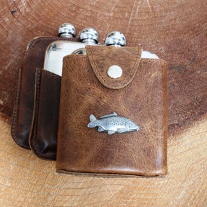 Könnte beinhalten: Ein brauner Lederflaschenhalter mit einer silbernen Flasche und einem Zinnfisch-Design.