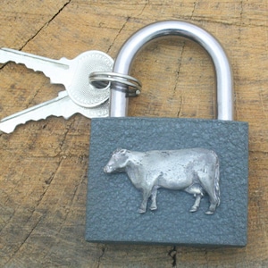 May include: A gray padlock with a silver cow design on the front. The padlock has a silver shackle and comes with two silver keys.