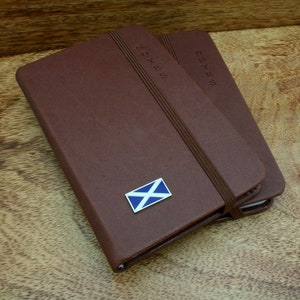 May include: Two brown leather notebooks with elastic closures. The top notebook has a small blue and white Scottish flag pin on the front cover. Both notebooks have the word "NOTES" embossed on the front cover.