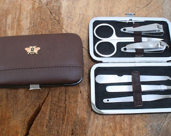 May include: A brown manicure set with a gold bee emblem on the case. The open case reveals stainless steel scissors, nail clippers, a nail file, a cuticle pusher, and tweezers. The set is on a wooden surface.
