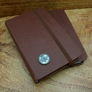 May include: Two brown leather bound notebooks with elastic closures. The top notebook has a silver metal Masonic symbol on the front cover. Both notebooks have the word "NOTES" embossed on the front cover.