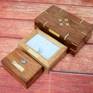 Peut inclure: Trois boîtes en bois avec des accents en laiton. La plus grande boîte a un motif décoratif en laiton sur le couvercle. La boîte du milieu a un cadre photo avec une photo en noir et blanc d'un bateau. La plus petite boîte a une figurine de cygne argentée sur le couvercle.