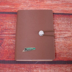Może przedstawiać: Brązowy notes w skórzanej oprawie ze srebrnym zapięciem i zieloną emaliowaną szpilką z pociągiem. Notes leży na czerwonym drewnianym tle.
