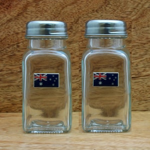 Könnte beinhalten: Zwei klare Glassalz- und Pfefferstreuer mit silbernen Deckeln. Jeder Streuer hat einen kleinen australischen Flaggenaufkleber auf der Vorderseite.