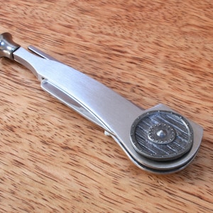 May include: A silver pipe tool with a shield-shaped design on the side. The tool has a small, round, silver cap on the end.