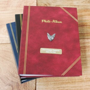 May include: Three photo albums stacked on a wooden surface. The top album is red with gold accents and the text "Photo Album" in gold lettering. A silver fox head is attached to the front cover. The text "Your message engraved here for free" is printed on a gold tag.
