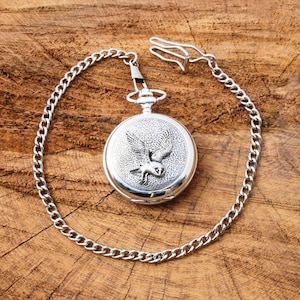 Könnte beinhalten: Silberne Taschenuhr mit Kette. Das Zifferblatt der Uhr zeigt ein gravierter Bild eines fliegenden Eulen.