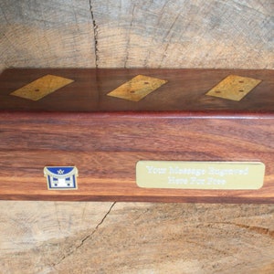 Pode incluir: Uma caixa de madeira com uma placa dourada que diz "Your Message Engraved Here For Free". A caixa tem três incrustações douradas com dois pontos cada. Há um pequeno emblema dourado e azul na frente da caixa.