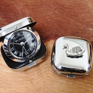 May include: A silver travel alarm clock with a black face and Roman numeral markings. The clock is open and the lid is beside it. The lid has a silver swan design.