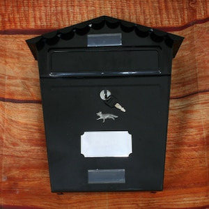 May include: Black metal mailbox with a peaked roof and a decorative fox emblem. The mailbox has a key and a silver nameplate. The mailbox is mounted on a wooden surface.