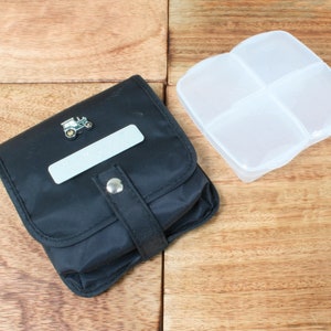 May include: A black zippered pouch with a small tractor pin and a white label. A clear plastic pill box with four compartments.