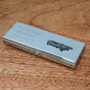 Könnte beinhalten: Eine silberne Metallbox mit einem Scharnierdeckel. Die Box hat eine gravierte Nachricht mit dem Text "Your Message Engraved Here." Die Box hat auch ein kleines silbernes Metalldesign mit einem Paar gekreuzter Gewehre auf dem Deckel.