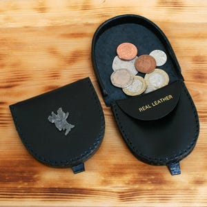 Pode incluir: Duas carteiras de moedas de couro preto, uma aberta e cheia de moedas. A carteira aberta tem as palavras "REAL LEATHER" impressas na aba. A outra carteira está fechada e tem um emblema prateado na frente.