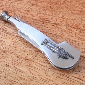 Può includere: Un attrezzo per pipa argentato con un design decorativo di due fucili. L'attrezzo ha una forma curva con una coppa a un'estremità e un pressino all'altra. Lo sfondo è una superficie di legno.