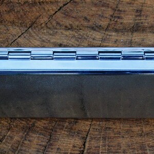 May include: A silver metal cigarette rolling tray with a hinged lid. The tray is closed and sitting on a wooden surface.