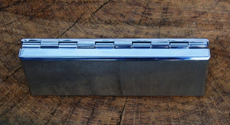May include: A silver metal cigarette rolling tray with a hinged lid. The tray is closed and sitting on a wooden surface.