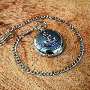 May include: A silver pocket watch with a black face and a blue and silver Royal Navy emblem. The watch is attached to a silver chain.