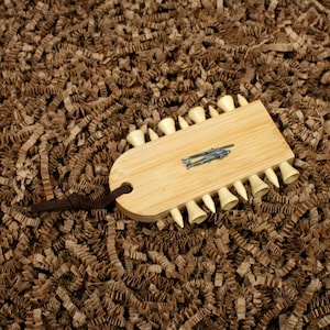 Könnte beinhalten: Ein hellfarbener Golf-Tee-Halter aus Holz mit einem dunkelbraunen Lederriemen. Der Halter hat einen kleinen Metallakzent und ist mit Golf-Tees gefüllt. Der Halter liegt auf einem Bett aus braunem Schredderpapier.