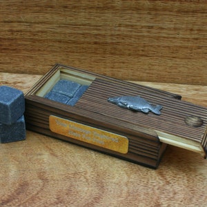 May include: A wooden box with a sliding lid, featuring a silver fish design. The box contains a set of gray whiskey stones. The box has a gold plaque with the text "Your Message Engraved Here For Free".