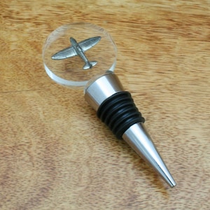May include: A silver bottle stopper with a clear glass top featuring a miniature silver aeroplane. The stopper has a black rubber ring and a pointed tip.