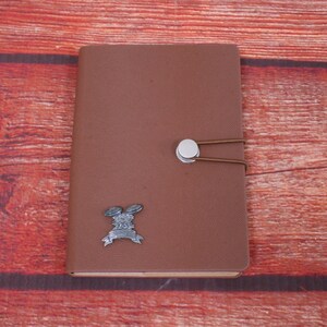 May include: A brown leather-bound journal with a silver clasp and a metal emblem of a pair of drumsticks with a ribbon and the number 25 on it.