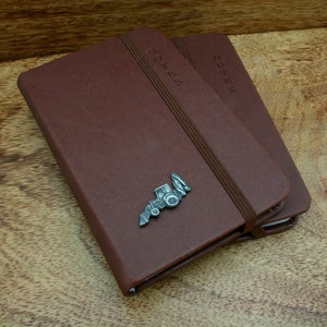 May include: Two brown leather bound notebooks with elastic closures. The front cover of the top notebook has a metal tractor charm attached. Both notebooks have the word "NOTES" embossed on the front cover.