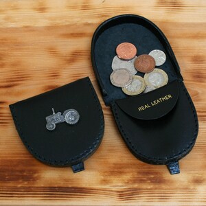 May include: Black leather coin purse with a silver tractor emblem. The purse is open, revealing several coins. The words "REAL LEATHER" are printed on the inside flap. The purse is on a wooden surface.