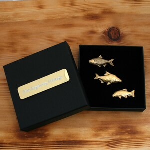 Peut inclure: Trois broches dorées en forme de poisson présentées dans une boîte noire ouverte avec un intérieur noir. Les broches sont visibles sur le fond sombre. Une plaque rectangulaire dorée avec le texte "Free Engraving Message" est sur la boîte.